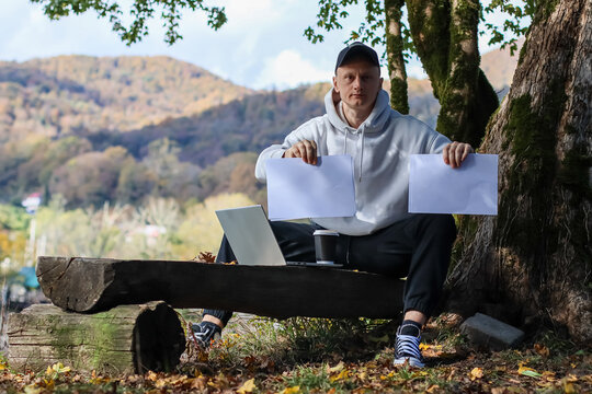 Young man with cap and hoodie sitting on log holding two blank papers with laptop outdoors. Man in casual wear outdoors, presenting pair of empty sheets, laptop on log in autumn natural setting