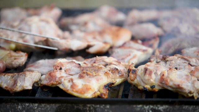 Meat roasting on grill close up, air flickering of heat and smoke rises carried by breeze, master with tongs turn pieces of meat out of focus