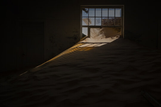 View of an abandoned room filled with desert sand dunes and sunlight streaming through a broken window in Kolmanskop, Karas Region, Namibia.