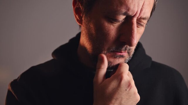 Portrait of a rugged man in dark hoodie, hand thoughtfully on chin, furrowed brow and closed eyes for worry, decision making, mental struggle concepts. 4K.