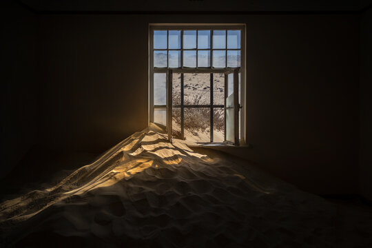 View of an abandoned house interior filled with a large sand dune under natural light through a window in Kolmanskop, Karas Region, Namibia.