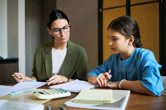 Middle aged woman tutoring schoolgirl, reviewing homework and explaining math at desk. Useful for education services, private tutoring, academic support, parenting campaigns