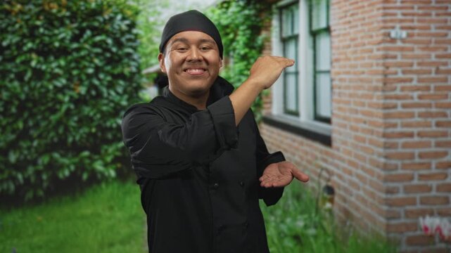 Man chef with palms up presenting at building entrance outdoors in black uniform and headscarf, smiling while offering empty hands; pride hospitality.