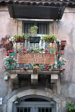 View of an ornate balcony decorated with colorful flowers and a wooden sign in the historic San Berillo district in Catania, Sicily, Italy.