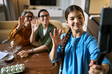 Obraz premium Teenage girl filming family baking video on smartphone, waving with mother and senior grandmother while preparing dough. Useful for family vlog, social media, parenting content marketing