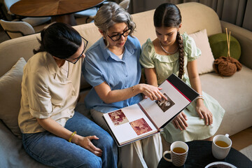 Senior woman with adult daughter and girl reviewing family photo album, sharing memories at home. Useful for genealogy, family heritage, generations, caregiving marketing, high angle shot
