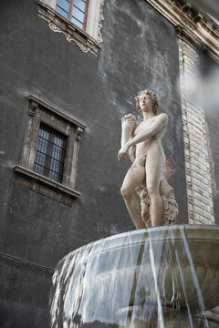 View of a marble statue on a historic fountain with water flowing in front of a dark stone building in Catania, Sicily, Italy.