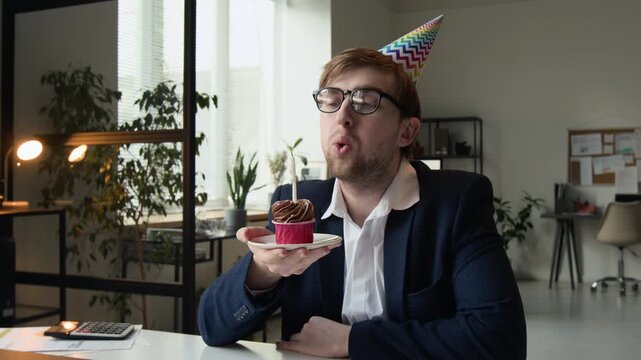 Portrait of indifferent Caucasian gen Z businessman dressed in white shirt and dark blue jacket holding plate with cupcake and blowing out candle on it at workplace
