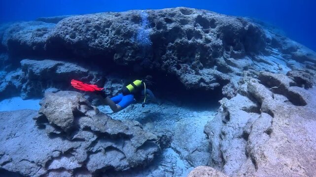 Scuba diver with pink fins exploring underwater cave entrance in the rocky seabed. Diver exploring the tunnel, vivid blue ocean. Sea cave in the stones, clear sea and swimming diver. 