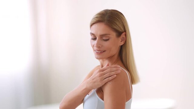 A woman with long blonde hair smiles while touching her shoulder in a well-lit indoor area. She is engaged in a self-care routine, appreciating her own beauty and wellness.