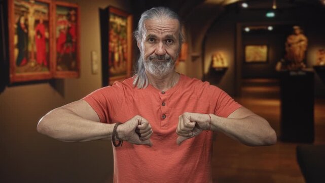 Man points both thumbs at his chest in a building art gallery with paintings and sculpture visible, wearing orange shirt; self critique skepticism.