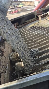 Side view of a concrete mixer truck discharging fresh material into a crane bucket at a sunny construction site.