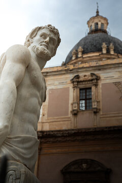 View of a white marble statue of a muscular man in front of the historic dome of a baroque church in Palermo, Sicily, Italy.