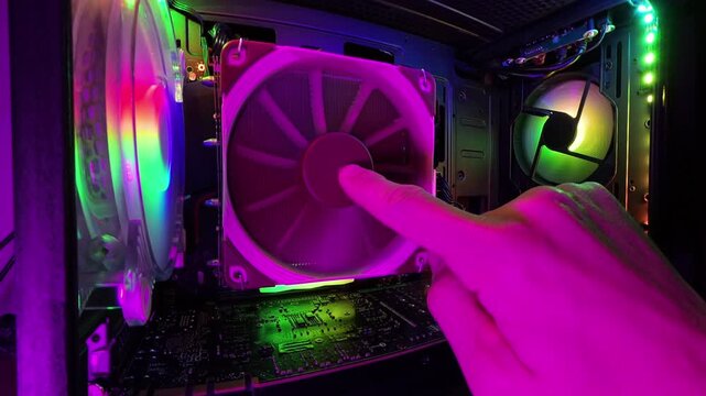 A zoomed-in view inside a gaming PC, featuring a hand stopping the RGB-lit cooling fan. The vibrant lighting and hardware components are illuminated in detail, creating a high-tech, immersive scene wi