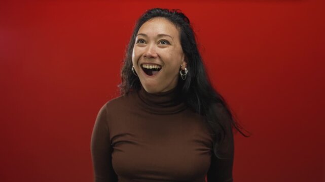 Woman with open mouth gasp and surprised gaze in studio on vivid red backdrop wearing brown turtleneck and hoop earrings; surprise joy energy.
