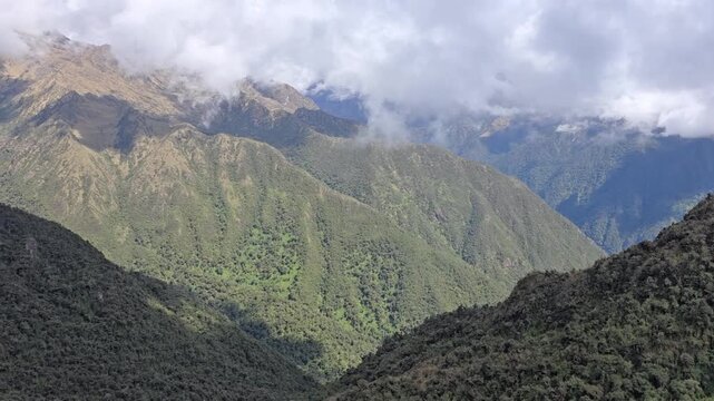 Cusco Region, Peru - 26 June 2024. Andes mountains along the Inca Trail with steep ridges, patchy vegetation, and low clouds covering upper peaks while sunlight lights the slopes.