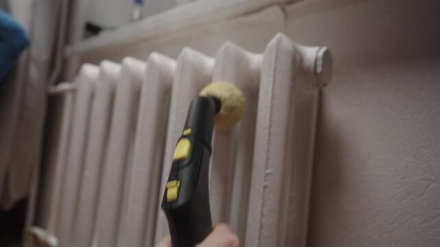 Hand using steamer with brush attachment for thorough cleaning of intricate radiator fins, ensuring household hygiene and effective maintenance of heating systems at home, close-up, slow motion.