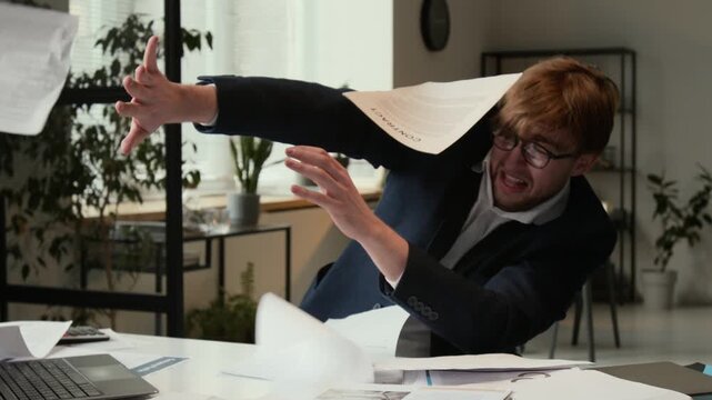 Zoom in shot of Caucasian frantic gen Z businessman in formalwear trying to avoid flying papers while typing on laptop at workplace