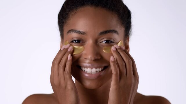 A woman with dark skin is applying gold eye patches to her under-eyes, smiling at the camera. This scene takes place indoors, highlighting a self-care moment.