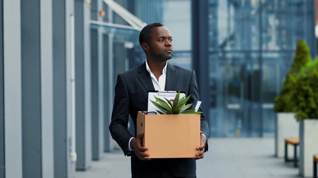 A man with dark skin stands outside an office building holding a box filled with personal items. He is dressed in business attire, reflecting a transition from work to new opportunities.
