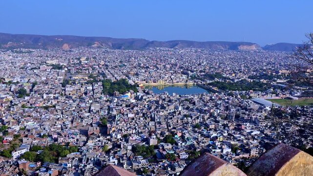 Daytime panoramic view of Jaipur city from Nahargarh Fort showing dense houses, Man Sagar Lake area, Aravalli hills in background, fort wall foreground and skyline of Jaipur.
