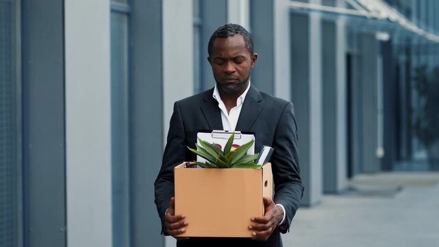 A man with dark skin stands outside an office building holding a box filled with files and a small plant. He appears thoughtful as he looks ahead while leaving the workplace.