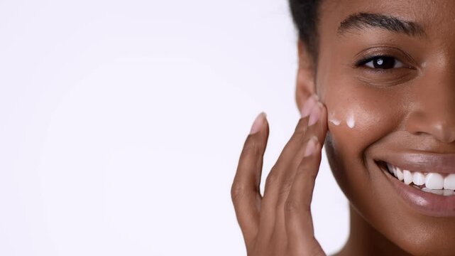 A woman with dark skin smiles while applying a white cream to her cheek in a bright and well-lit area. She showcases a positive expression while focusing on her skin care routine.