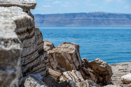 Layered salt and limestone rocks on the Jordanian shore of the Dead Sea with blue water and arid mountains in the background.