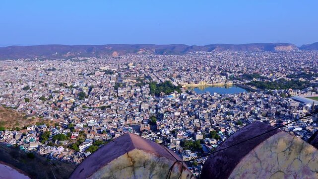Daytime panoramic view of Jaipur city from Nahargarh Fort showing Man Sagar Lake, dense urban houses, Aravalli hills in background, fort wall foreground and wide skyline of Jaipur Rajasthan India.