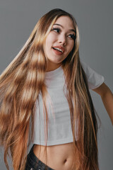 Young woman with long straight hair wearing white crop top and jeans posing with smile on gray...