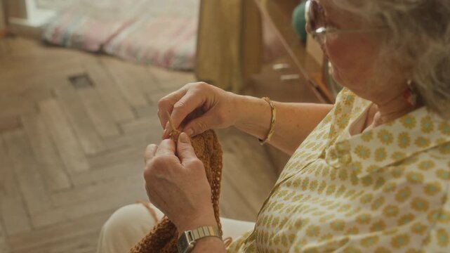 Over shoulder shot of elderly woman focused on knitting wool scarf with needles during relaxing leisure activity at home