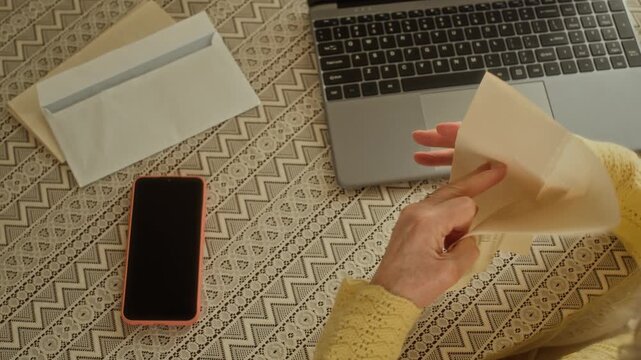 Top view of elderly woman opening envelope with utility payment statement comparing information with data on laptop screen while sitting at table with devices in kitchen