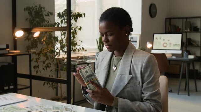 Tilt up view of joyful Black young woman taking bundle of dollar bills and smelling money with enjoyment during workday in office