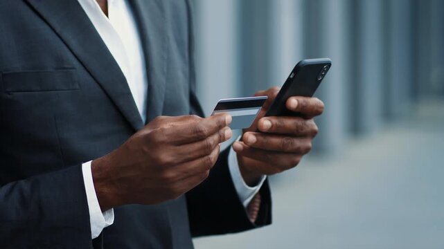 A businessman with dark skin stands outdoors, holding a smartphone in one hand and a credit card in the other. He is engaging in a mobile payment activity.
