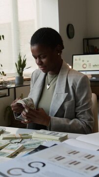 Vertical rack focus view of happy Black female young entrepreneur examining dollar bundles with excitement during workday in office
