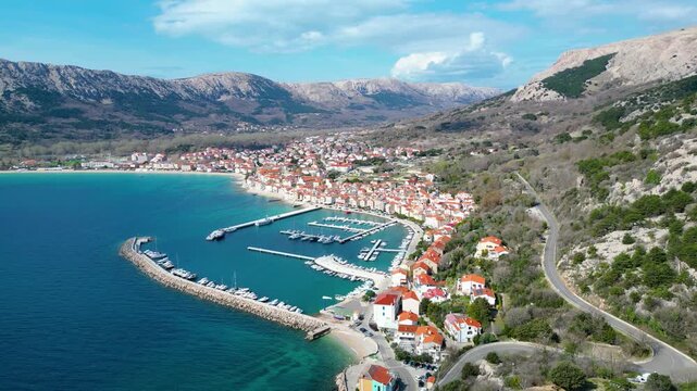 Stunning aerial view of Baska, Krk Island, Croatia. A picturesque coastal town with a vibrant marina, nestled against rugged mountains under a clear spring sky.