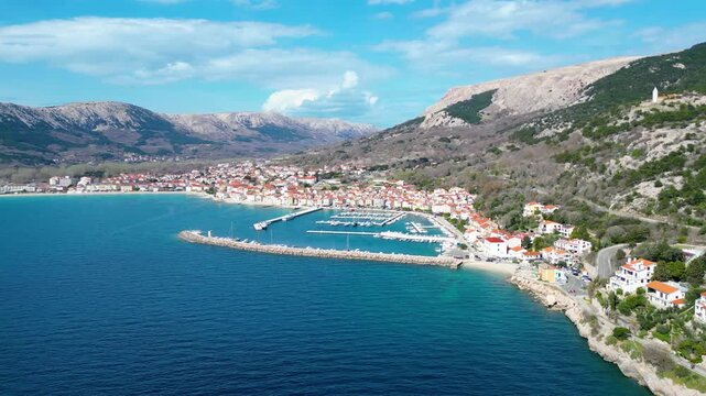 Stunning aerial view of Baska, Krk Island, Croatia. A picturesque coastal town with a vibrant marina, nestled against rugged mountains under a clear spring sky.