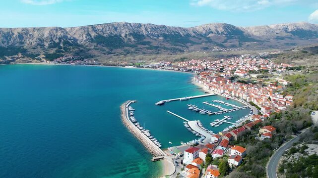 Aerial view of Baska's vibrant harbor and red-roofed town nestled against dramatic mountains on Krk Island, Croatia. Clear skies and turquoise waters.