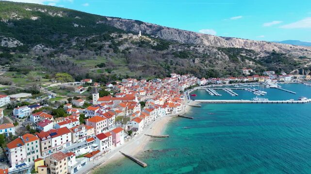 Aerial view of Baska, Krk Island, Croatia. Traditional red-roofed homes hug the turquoise Adriatic Sea, backed by rugged mountains. A picturesque early spring scene.