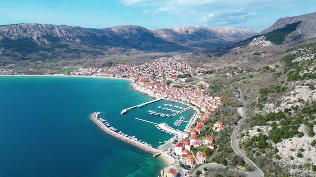 Aerial view of Baska, Krk Island, Croatia. A charming coastal town with red-tiled roofs, a bustling marina, and clear turquoise Adriatic waters against a rugged mountain backdrop.