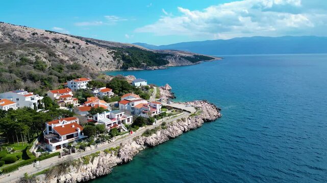 Aerial view of a charming Croatian coastal village on Krk Island, featuring red-roofed homes nestled on rocky cliffs overlooking the clear Adriatic Sea under a bright spring sky.