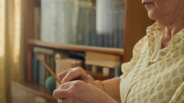 Tilt up shot of elderly woman wearing glasses knitting wool sweater using needles while enjoying peaceful hobby at home