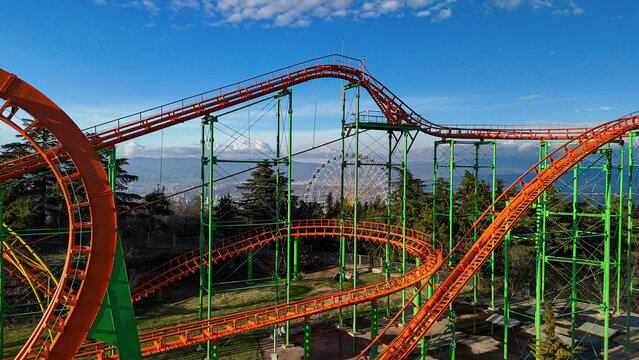 Orange rollercoaster tracks and green supports twist through mtatsminda park, racing past the ferris wheel with tbilisi city and mountain views under a bright blue sky