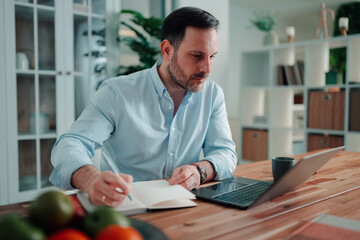 Fototapeta premium Man focused on work, writing in a planner while using a laptop on a wooden desk. Concentrating on remote tasks