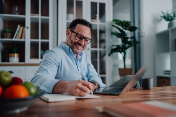 Fototapeta premium Mature man working from home, writing notes and smiling while using laptop for remote business