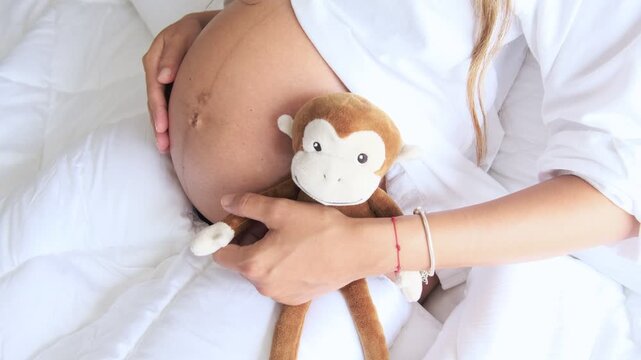 Pregnant woman lying on a white bed, gently caressing her belly and holding a soft monkey toy