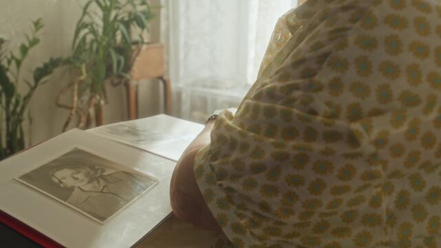 Tilt up shot of elderly woman with white hair smiling while flipping pages of retro photo album reminiscing over past standing in sunlit living room