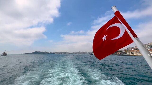 Red Turkish national flag blowing in wind on ferry with visible sea wake and Istanbul waterfront in background under cloudy sky