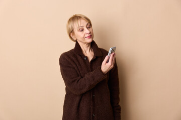 Fototapeta premium Mature woman in brown coat looks at smartphone with slight smile on beige background. Concept of technology, communication, and casual lifestyle for middle aged people.