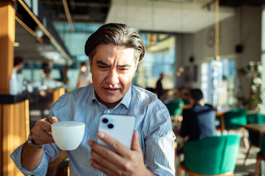 Middle-aged man drinking coffee and using smartphone in modern cafe
